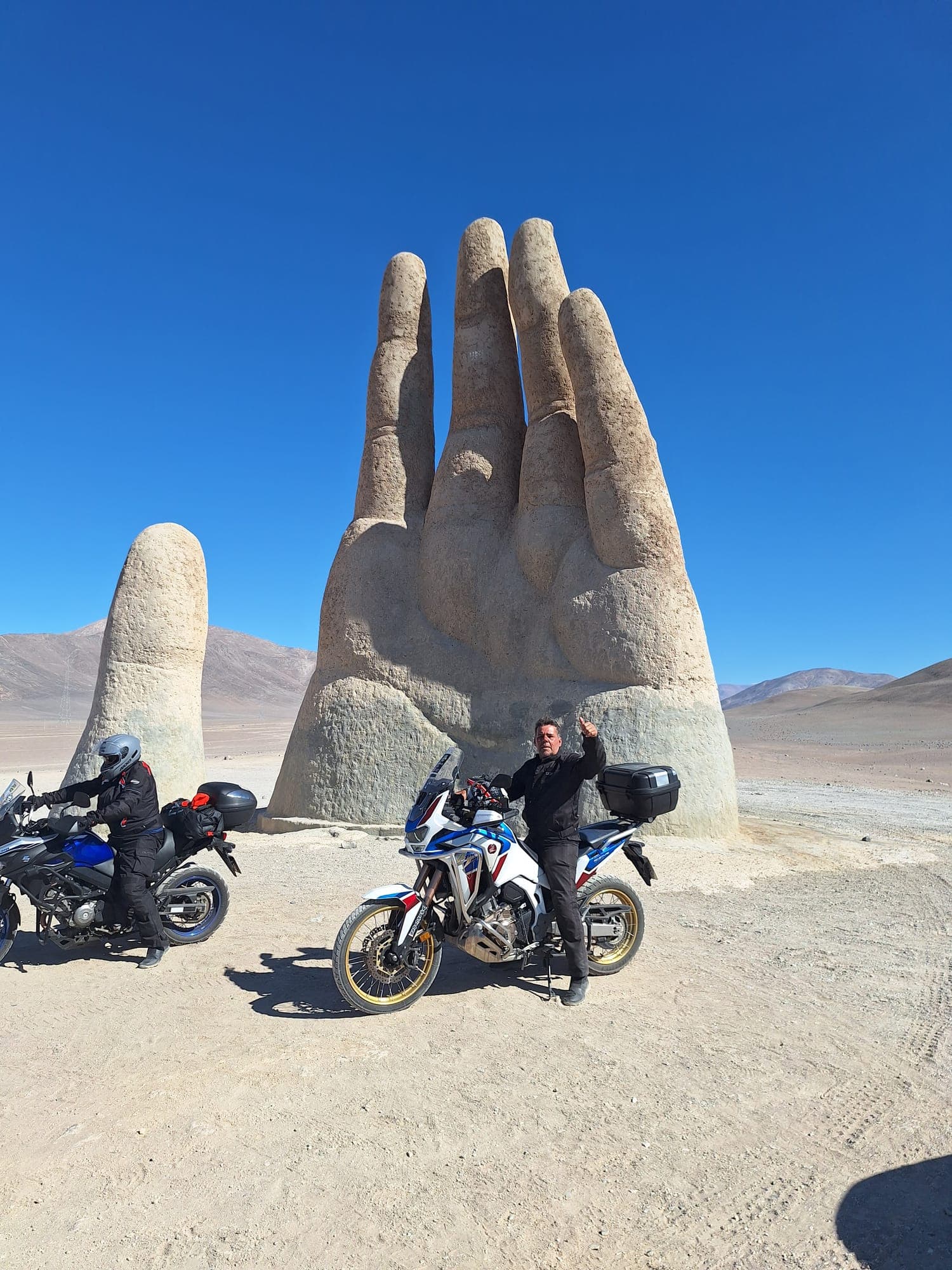 Duas motos de aventura e um piloto diante da Mano del Desierto, a mão gigante no deserto, sob céu azul