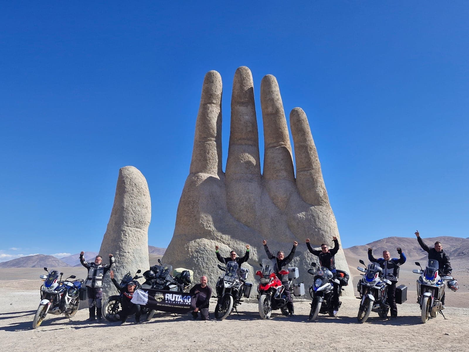 O grupo inteiro comemorando de braços erguidos ao lado das motos, diante da Mano del Desierto
