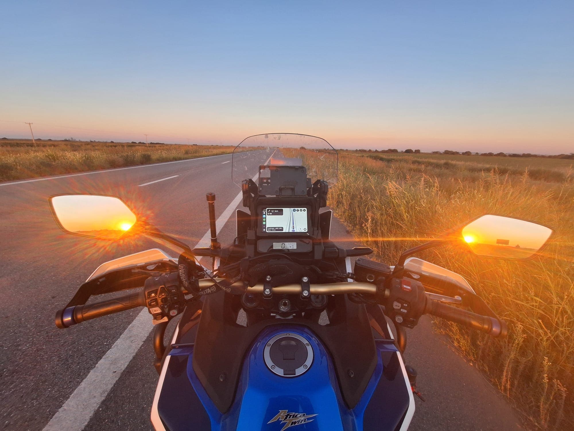 Guidão de uma moto com os piscas acesos, de frente para o pôr do sol na estrada